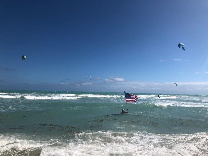 Glen Bradford in the ocean holding an American flag with kites flying overhead